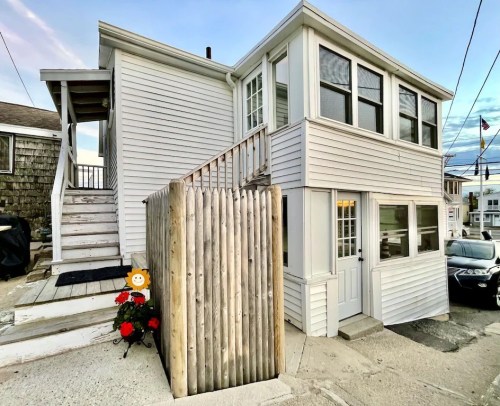 A white two-story house with a wooden fence, stairs, and flower pots in front, set against a clear sky.