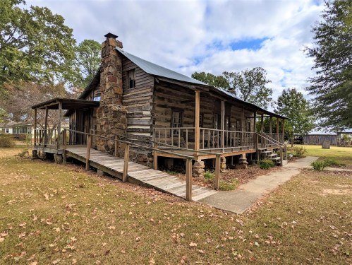 A rustic wooden cabin with a stone chimney, surrounded by trees and a grassy area under a partly cloudy sky.