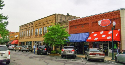 A street view of shops with colorful awnings, parked cars, and trees lining the sidewalk on a cloudy day.