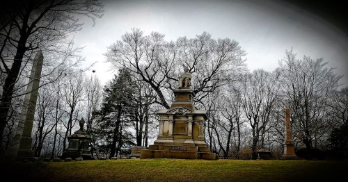 A somber cemetery scene featuring ornate gravestones and bare trees under a cloudy sky.
