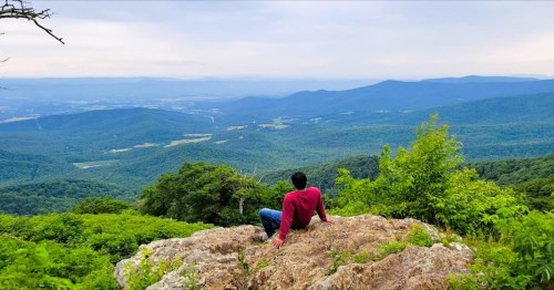 A person sits on a rock, gazing at a vast green valley and distant mountains under a cloudy sky.