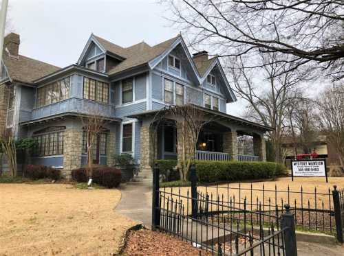 A large blue Victorian-style house with a stone foundation and a for-sale sign in the front yard.