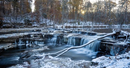 A serene winter scene featuring a waterfall surrounded by snow-covered rocks and trees, with a calm river below.