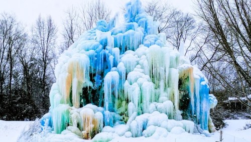 A colorful ice formation with cascading layers of blue, green, and orange, surrounded by bare trees in winter.