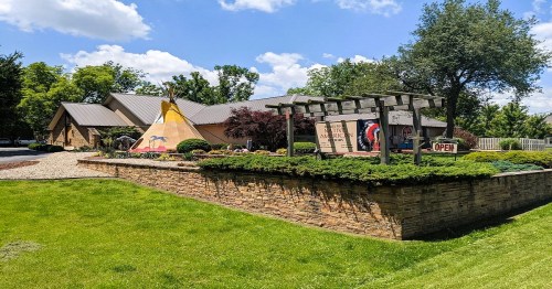 A colorful teepee stands beside a modern building with a landscaped garden and an "Open" sign.