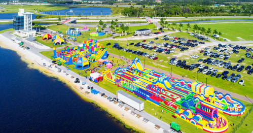 Aerial view of a colorful inflatable water park beside a lake, with cars parked nearby and palm trees in the background.