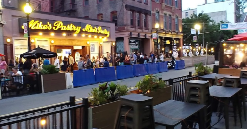 A busy street scene with a long line outside a pastry shop, surrounded by outdoor seating and city buildings.