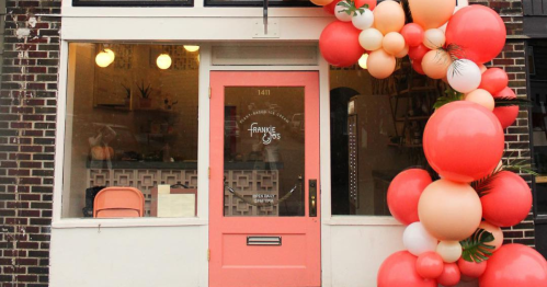 A storefront with a coral door and a colorful balloon arch in front, featuring the name "Frankie" on the window.