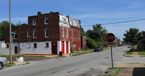 A red-brick building on a street with a stop sign, surrounded by greenery and clear blue skies.