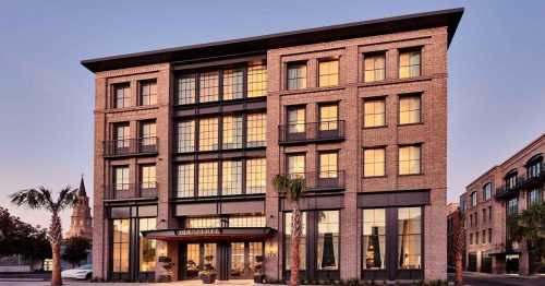 A modern brick hotel building with large windows, palm trees, and a historic church in the background at sunset.