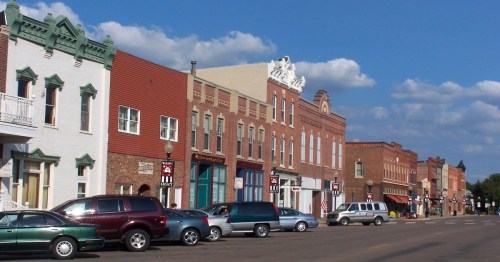 A quaint street lined with historic buildings and parked cars under a blue sky with fluffy clouds.