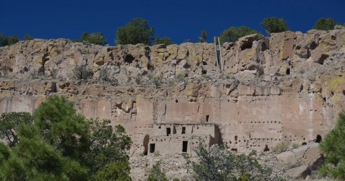 Ancient cliff dwellings nestled in a rocky landscape, surrounded by greenery and a clear blue sky.