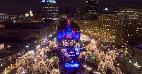 Aerial view of a city square at night, featuring a lit-up building and trees adorned with colorful lights.