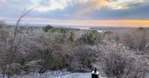 A winter landscape with frosted trees and a serene view of a lake at sunrise, under a cloudy sky.