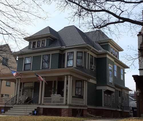 A large, green Victorian-style house with a porch, featuring two American flags and multiple windows.