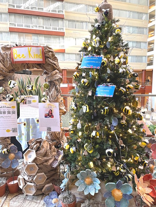 A decorated Christmas tree with bee-themed ornaments and a "Bee Kind" sign, surrounded by flower displays.