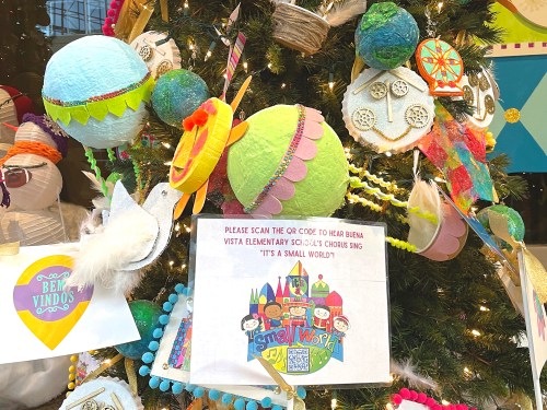 Colorful ornaments and decorations on a Christmas tree, with a sign inviting viewers to scan a QR code for a chorus performance.