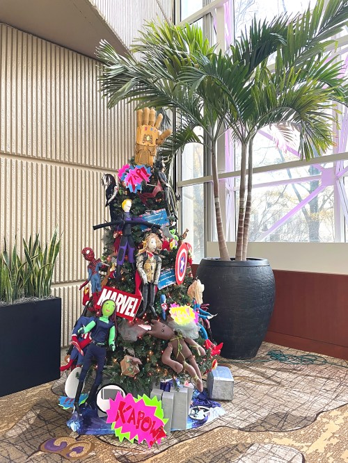 A colorful Christmas tree decorated with superhero toys, comic book elements, and vibrant signs in a modern indoor setting.