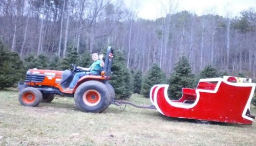 A boy sits on a tractor pulling a red sleigh through a Christmas tree farm. Trees are visible in the background.