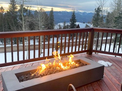 A modern fire pit on a wooden deck, surrounded by snow-covered trees and mountains under a sunset sky.