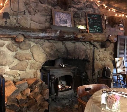 Cozy interior featuring a stone fireplace, wooden beams, and rustic decor with a stack of firewood nearby.