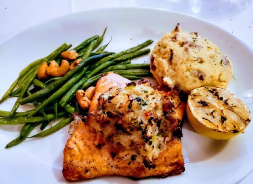 A plate featuring baked salmon topped with herbs, green beans, mashed potatoes, cashews, and a lemon wedge.