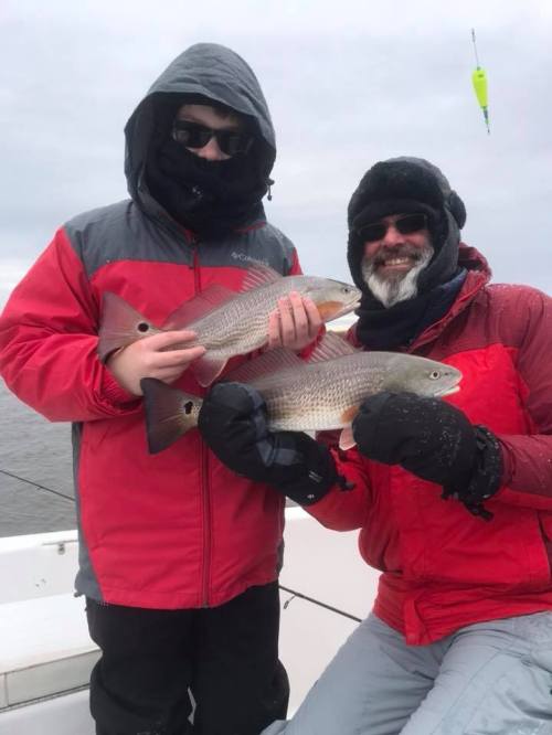 Two people in winter gear hold up large fish while on a boat, with overcast skies in the background.