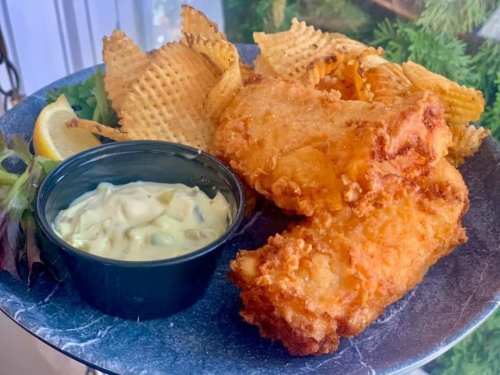 A plate of crispy fried fish fillets with waffle fries and a side of tartar sauce, garnished with a lemon wedge.
