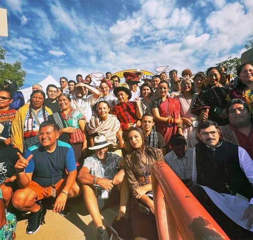 A diverse group of people poses together outdoors, smiling, with colorful clothing and a blue sky in the background.