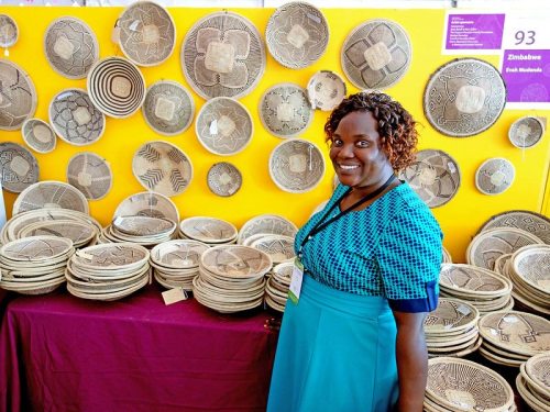 A woman in a blue dress stands beside a display of intricately designed woven baskets on a yellow background.