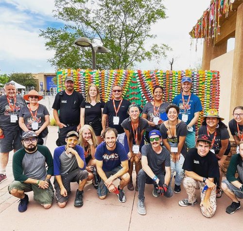 A diverse group of people poses together in front of a colorful wall at an outdoor event, smiling and wearing casual attire.