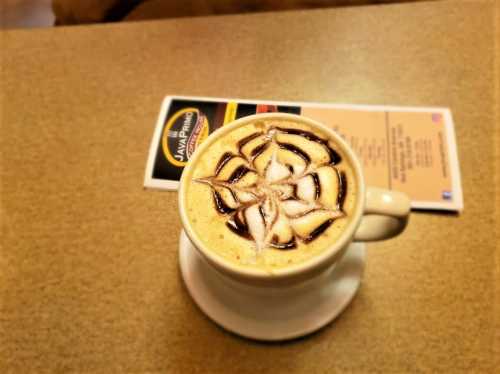 A cup of coffee with a decorative foam design on top, placed on a table with a menu in the background.