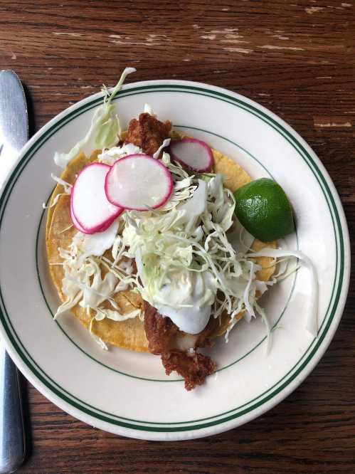 A plate with a taco topped with fried chicken, shredded cabbage, radishes, and a lime wedge on a wooden table.