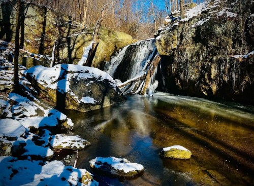A serene winter scene featuring a waterfall surrounded by snow-covered rocks and trees under a clear blue sky.