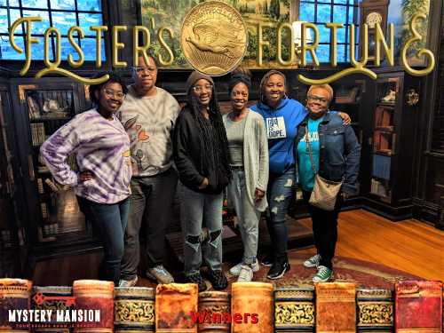 A group of six smiling individuals stands together in a cozy room, celebrating their win at "Fosters of Fortune."