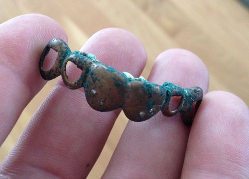 A hand holding a small, weathered bronze artifact with green patina, featuring circular cutouts along its length.