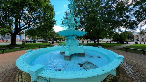 A turquoise fountain surrounded by trees and benches in a park, with a clear blue sky above.