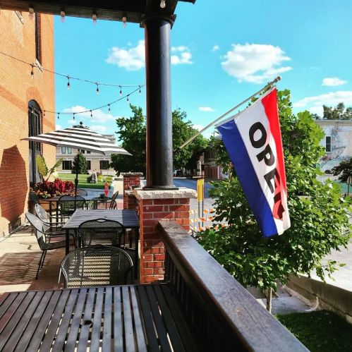 A sunny outdoor patio with tables, a red, white, and blue "OPEN" flag, and greenery in the background.