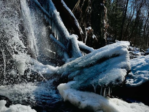A frozen waterfall with ice formations and flowing water, surrounded by trees in a winter landscape.