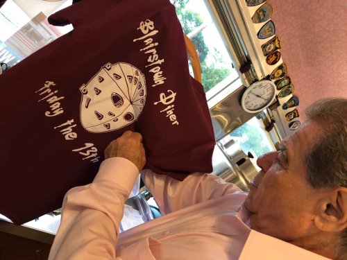 A person holds up a maroon t-shirt featuring a graphic of a hockey mask and the text "Blain's Diner, Friday the 13th."