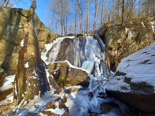 A frozen waterfall surrounded by snow-covered rocks and trees under a clear blue sky.