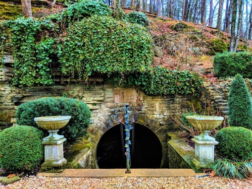 A stone entrance surrounded by greenery and decorative planters, leading into a dark tunnel in a wooded area.