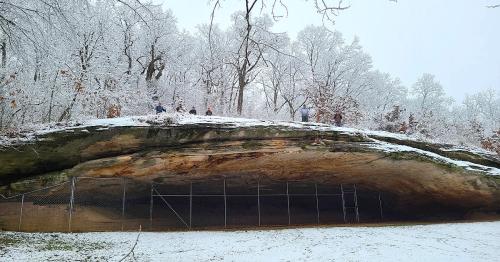 A snowy landscape featuring a large rock overhang with a fenced area below and people standing on top of the ledge.