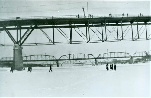Black and white photo of a bridge over a snowy landscape, with people walking on the ice below.