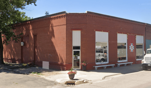 Red brick building with large windows, planters, and a sign featuring a logo, located on a street corner.