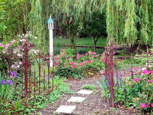 A lush garden with colorful flowers, a winding path, and a decorative white post surrounded by greenery and trees.