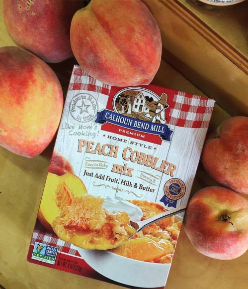 A box of Calhoun Bend Mill peach cobbler mix surrounded by fresh peaches on a wooden surface.