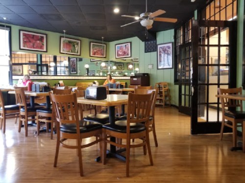 A cozy restaurant interior with wooden tables and chairs, featuring green walls and framed artwork. Two patrons are seated.