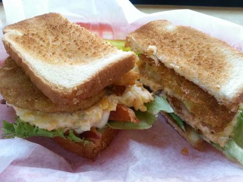 A toasted sandwich with lettuce, tomato, and a fried patty, cut in half and served on a red paper wrapper.
