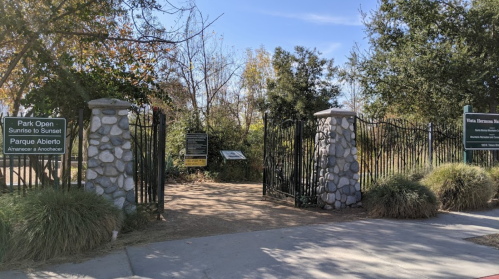 Entrance to a park with stone pillars and gates, surrounded by trees and signage about park hours and rules.